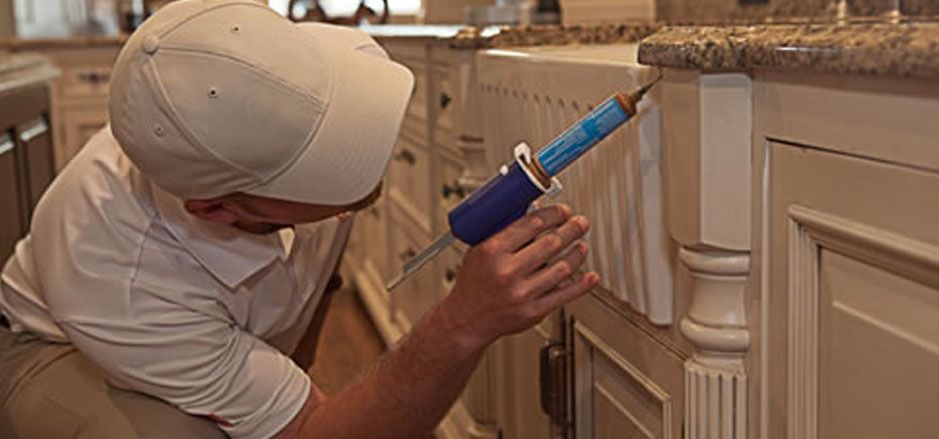 Person in a cap using a blue and black tool to apply a substance to a white kitchen cabinet. — Billy Lanes Pest Control Services in Kiama, NSW