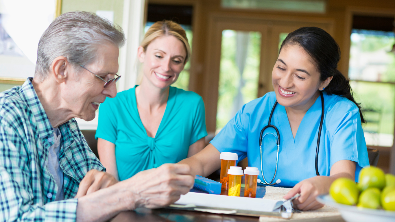 Senior man and two smiling women reviewing medicine bottles; one is a nurse.