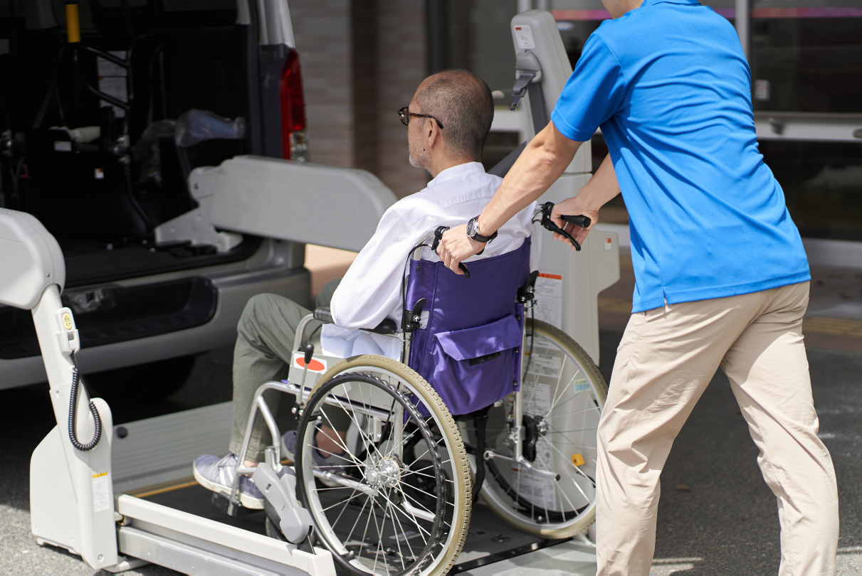 Elderly woman in glasses smiles at a person off-screen, seated at a table. Wheelchair present, living room setting.