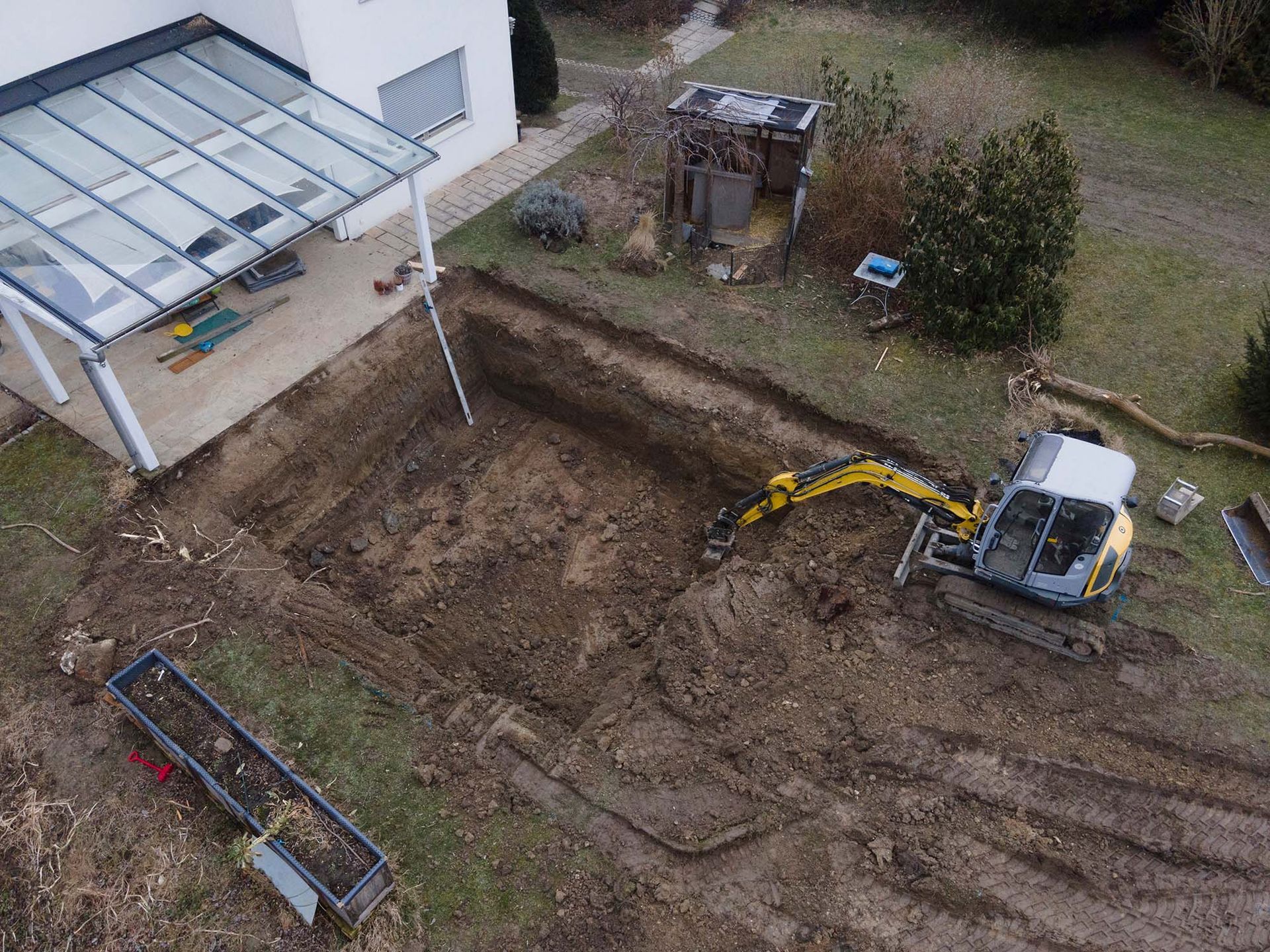 An excavator digging a trench in a muddy backyard next to a house with a patio.