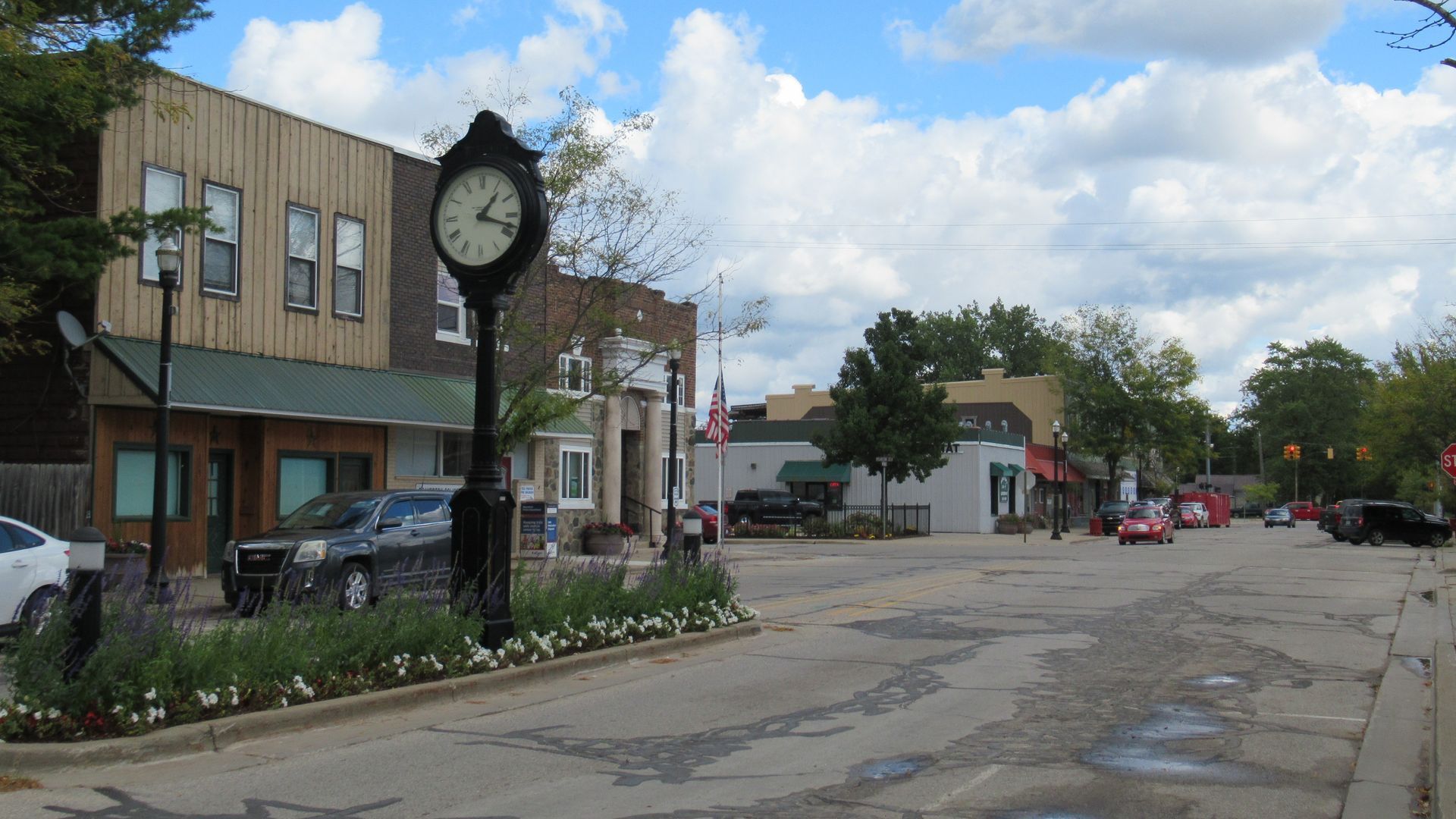 Street view with clock tower, buildings, cars, trees, and cloudy sky.