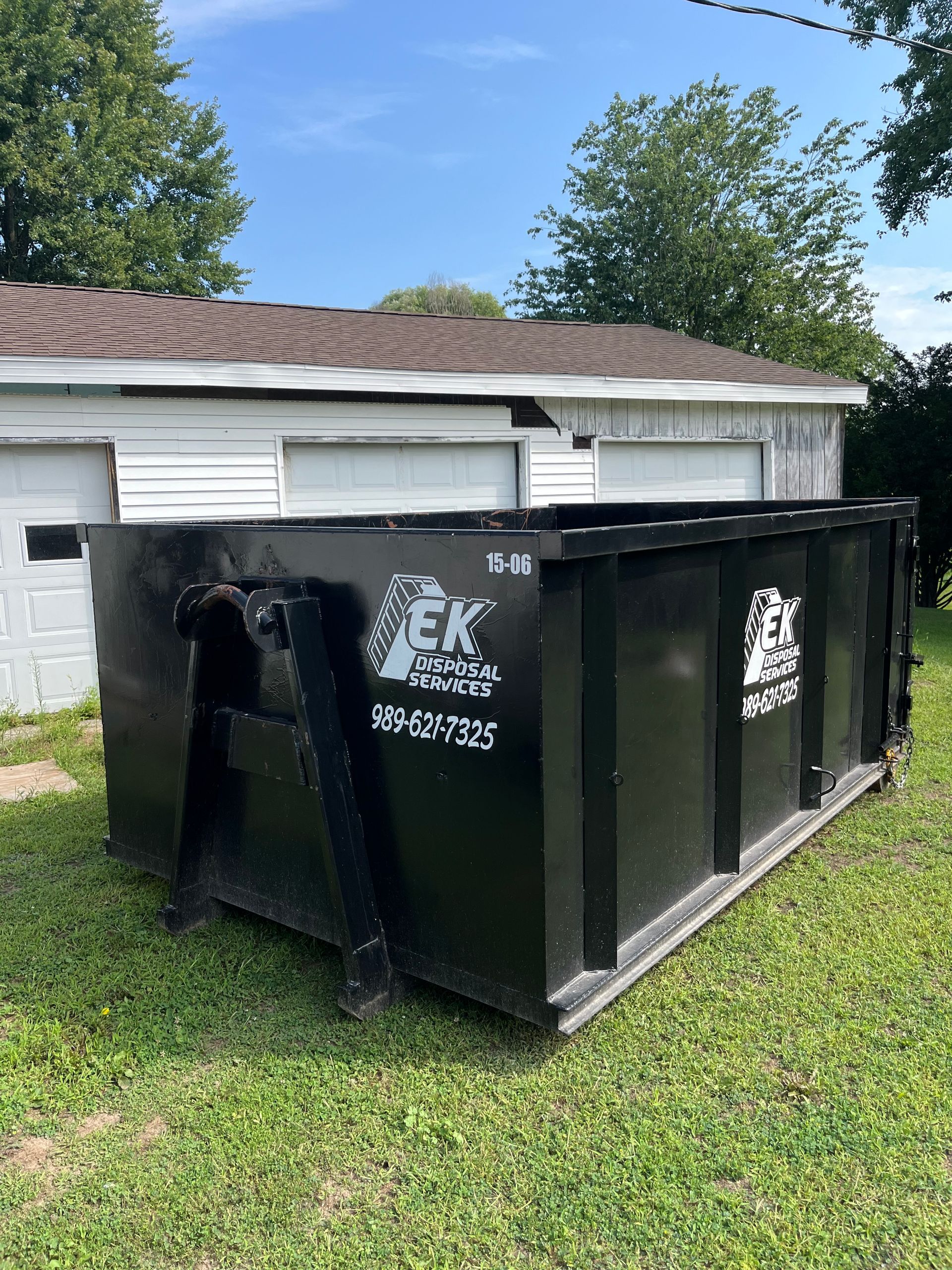 Gray dumpster filled with yard waste on a residential street in front of a two-story house.