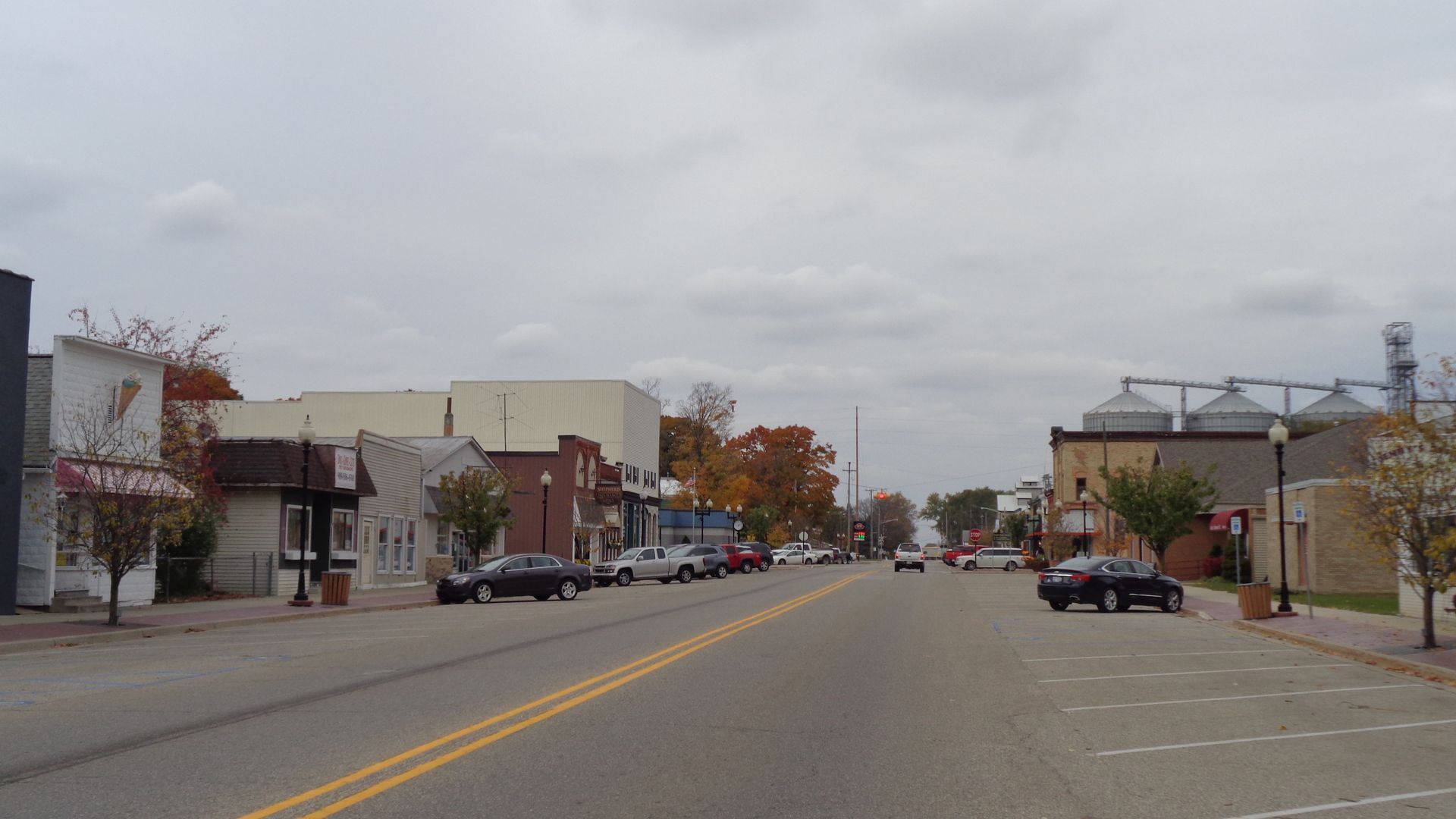 Street view of a small town with low buildings, cars, trees, and cloudy sky.