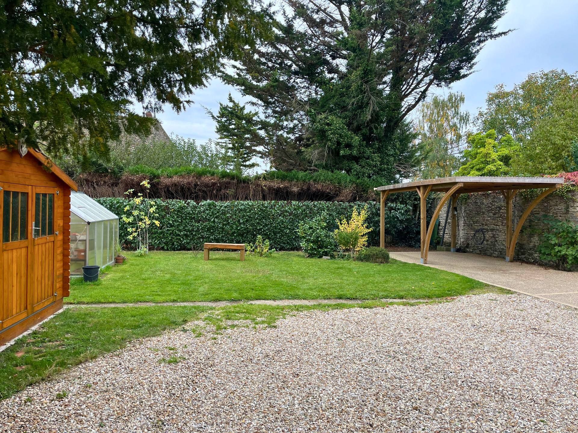 Une cour avec un petit abri en bois, une serre, une pelouse, un banc, un parking couvert, une allée en gravier et des arbres.