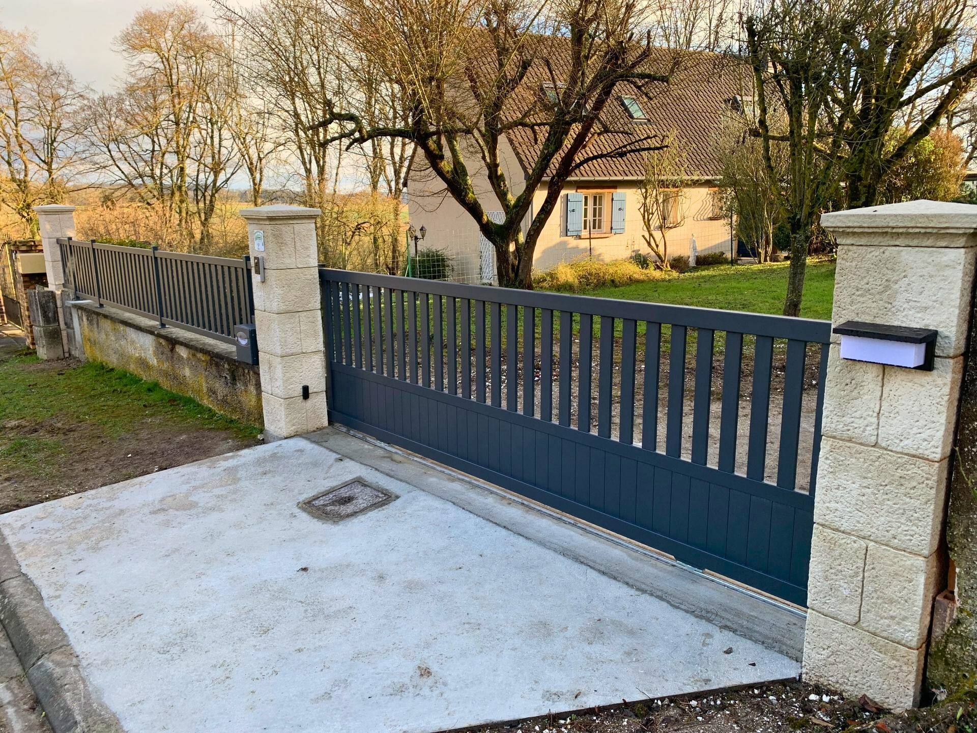 Portail en métal gris à barreaux verticaux, piliers en pierre, allée en béton et maison en arrière-plan.
