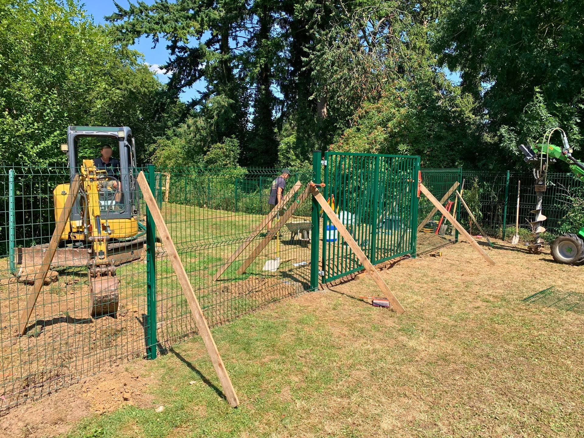 Chantier : Des ouvriers construisent une clôture métallique verte à l'aide d'une excavatrice dans une zone herbeuse, avec des arbres en arrière-plan.