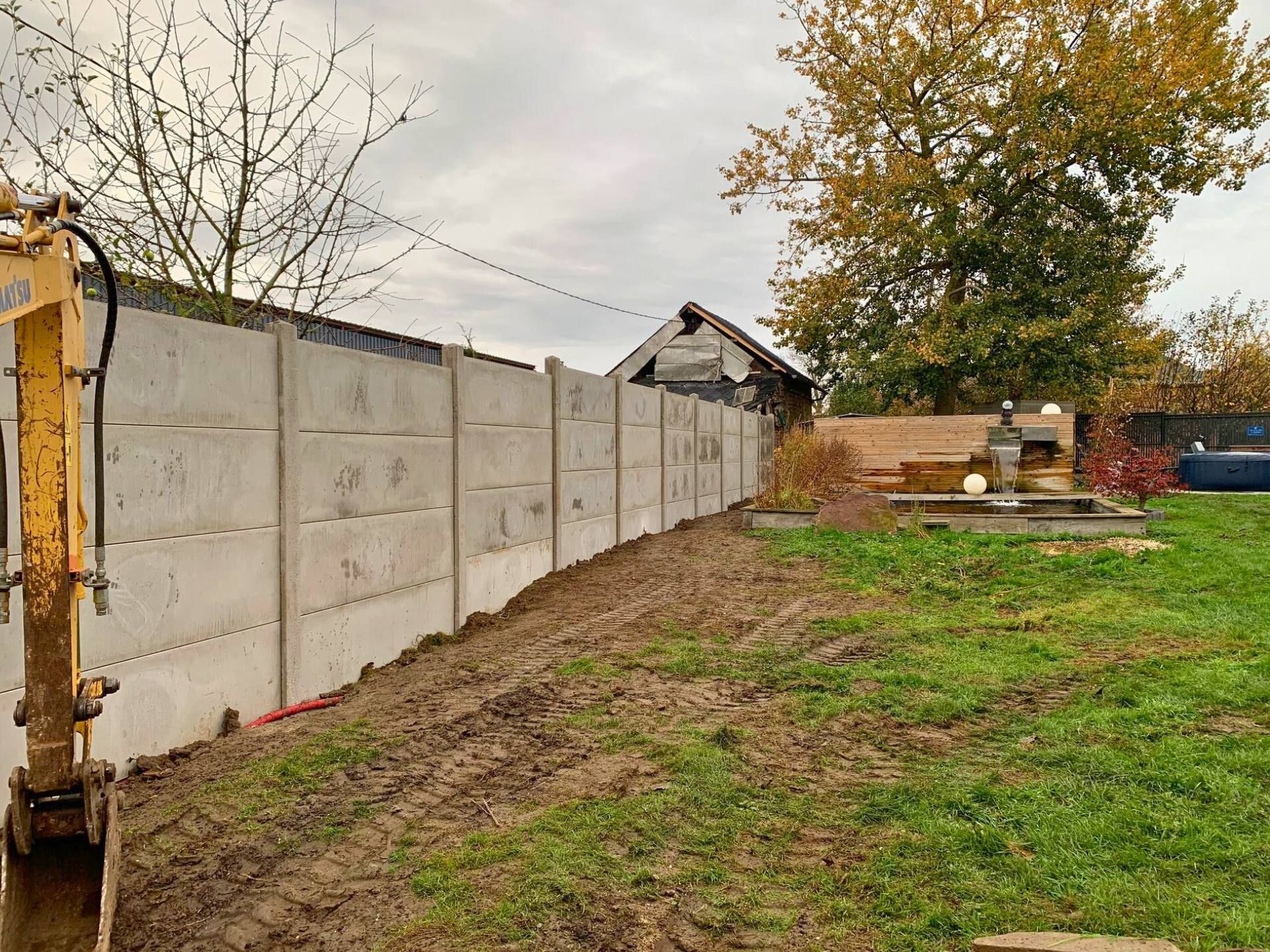Chantier de construction avec une clôture en béton en construction, une petite excavatrice à gauche et une cour arrière.