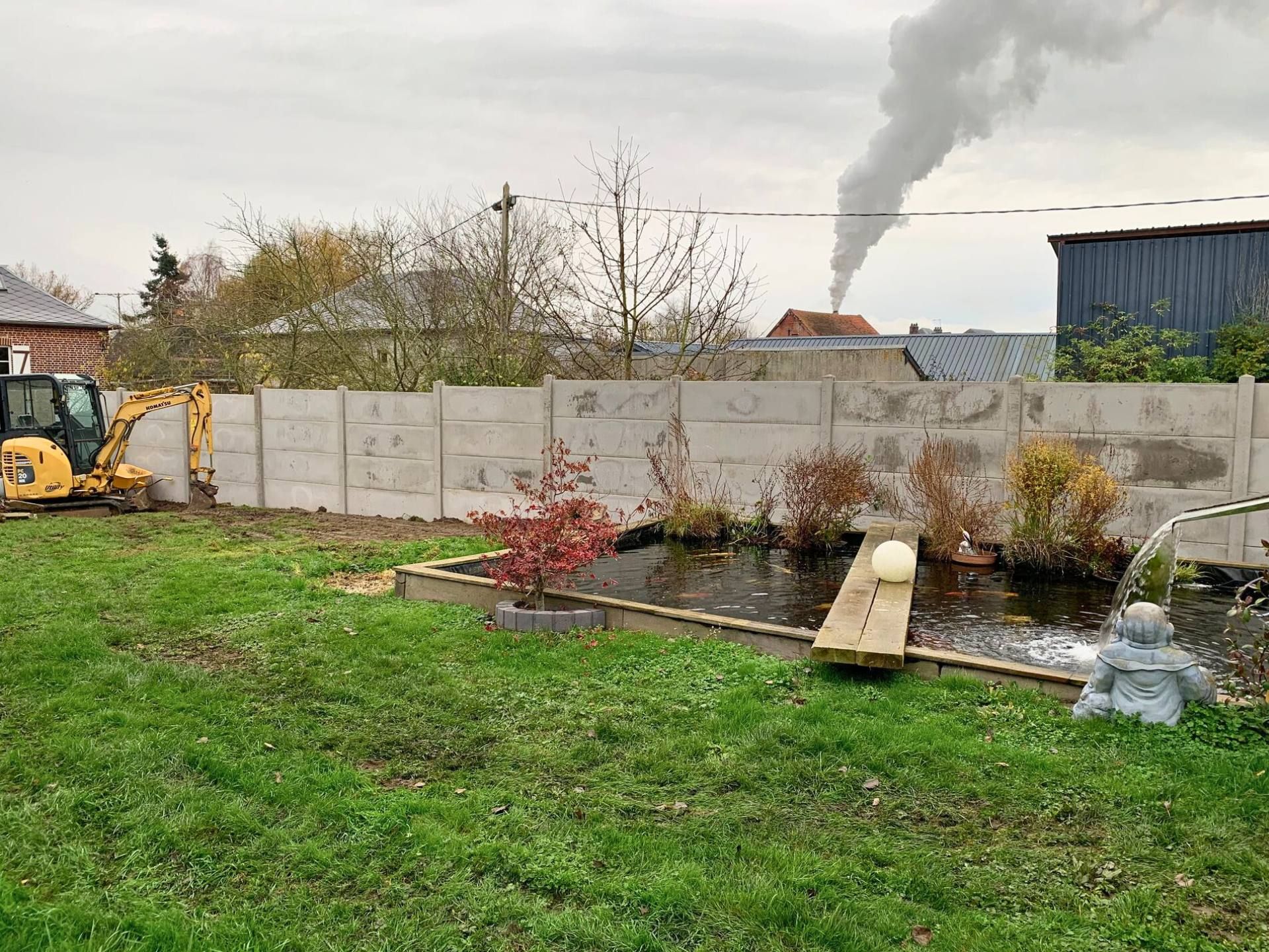 Une scène de jardin avec un étang, une clôture en béton et une petite pelleteuse. De la fumée s'échappe d'un bâtiment au loin.