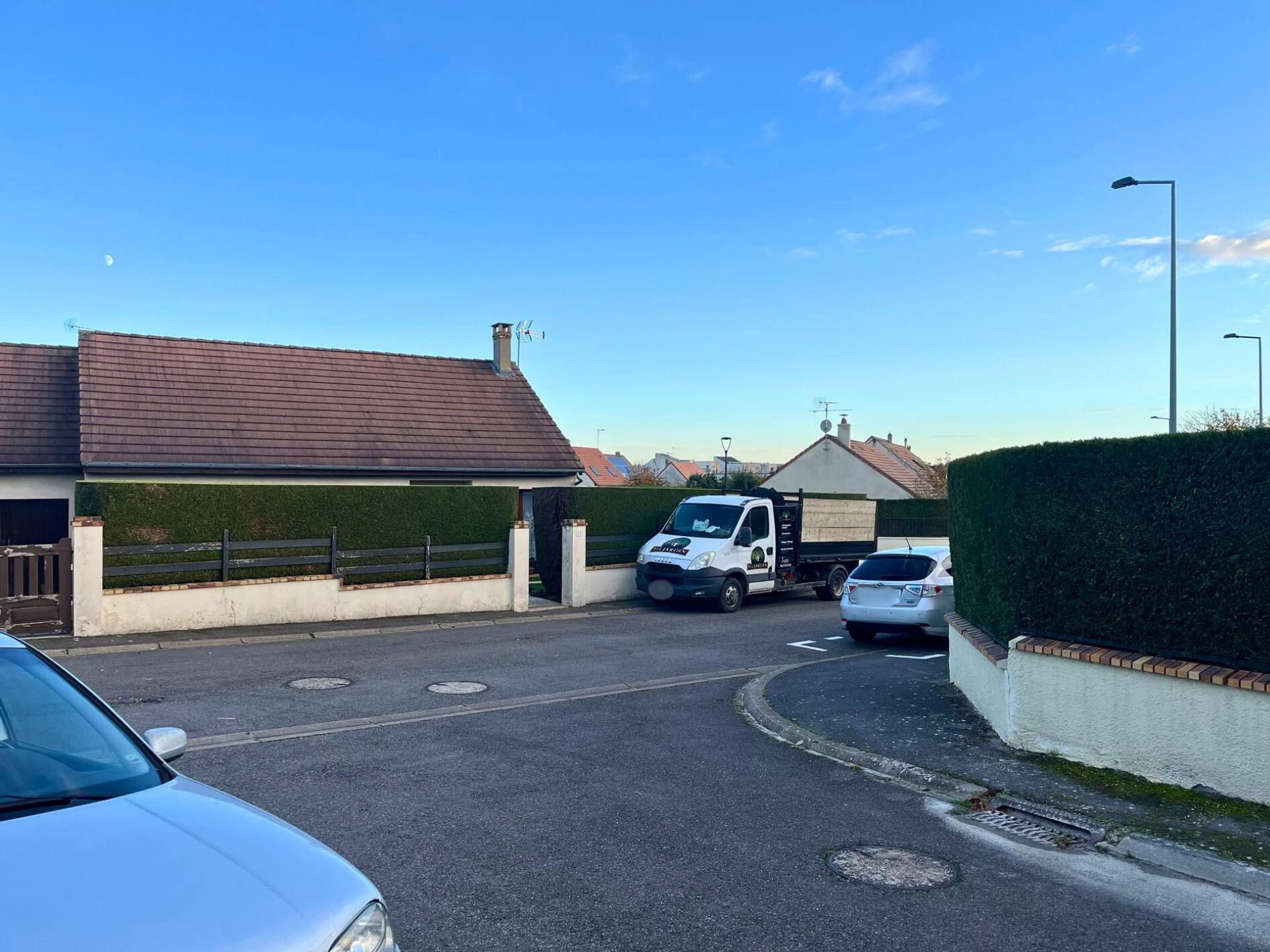 Un camion et une voiture sont garés dans une rue résidentielle, bordés de maisons et de haies vertes, sous un ciel dégagé.
