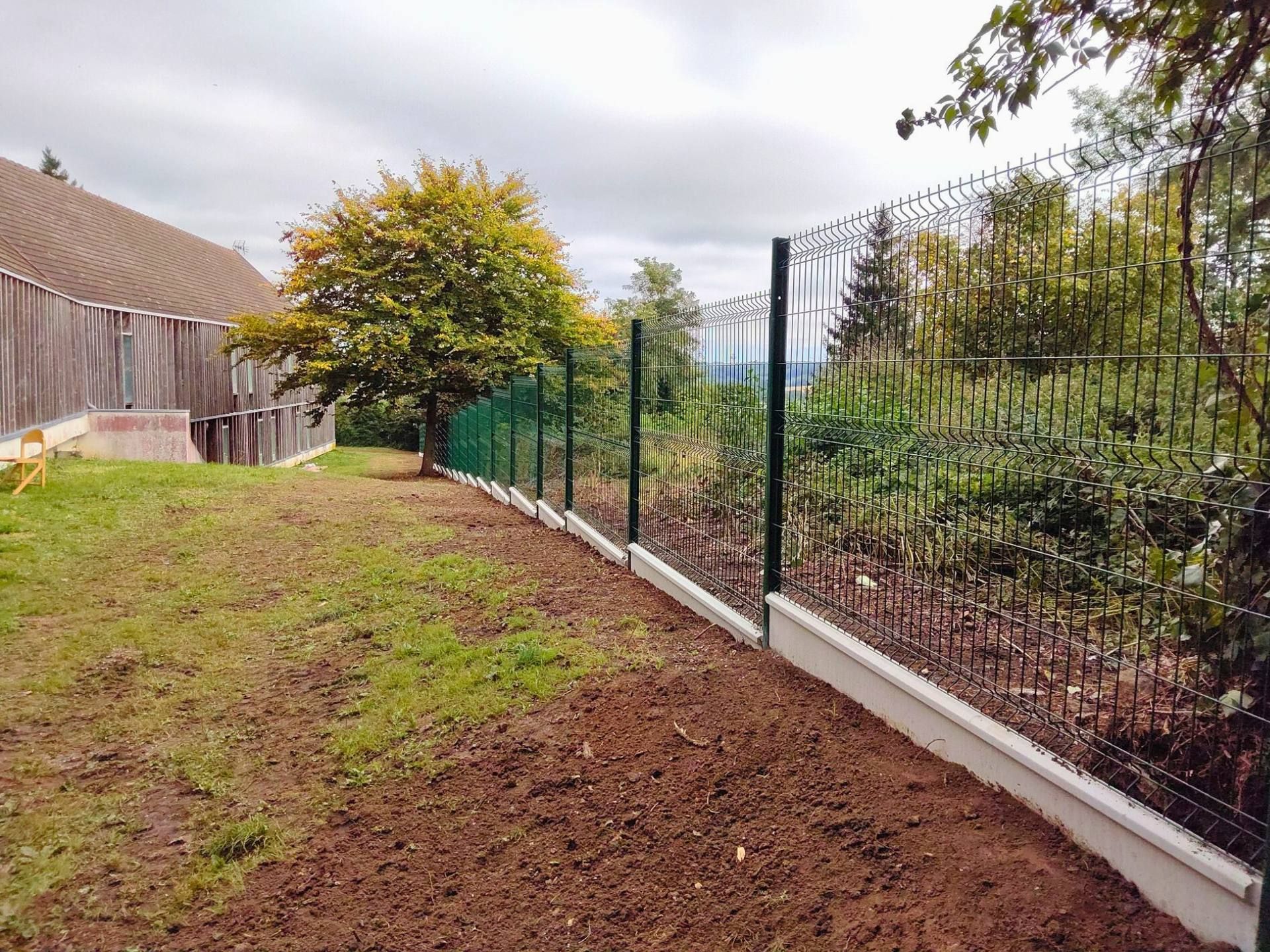 Clôture métallique verte sur socle en béton, délimitant un jardin. Terre brune et herbe verte. Ciel nuageux.