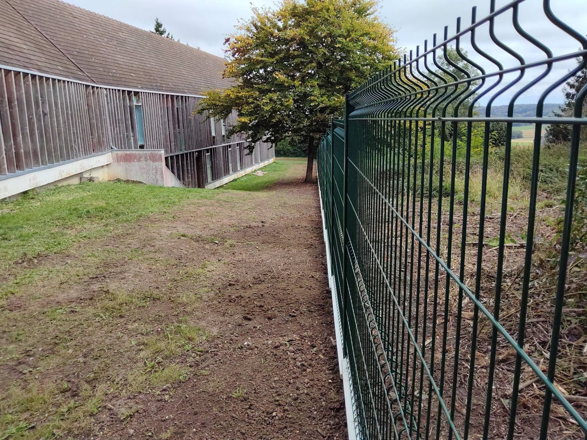 Clôture métallique verte le long d'un bâtiment, avec un arbre et de l'herbe entre les deux.