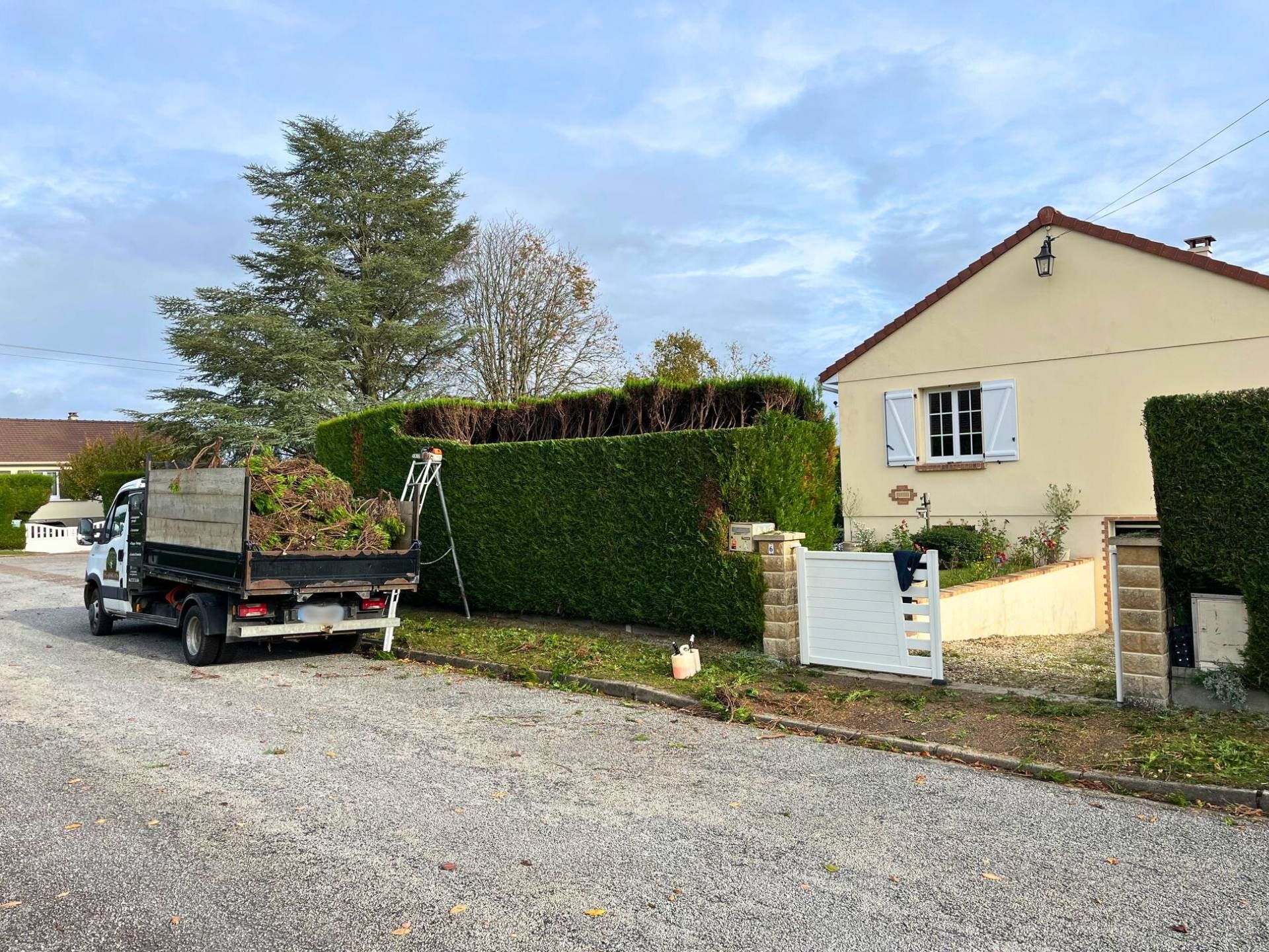 Un camion chargé de branches est stationné près d'une maison et d'une haie taillée, sur une allée de gravier.