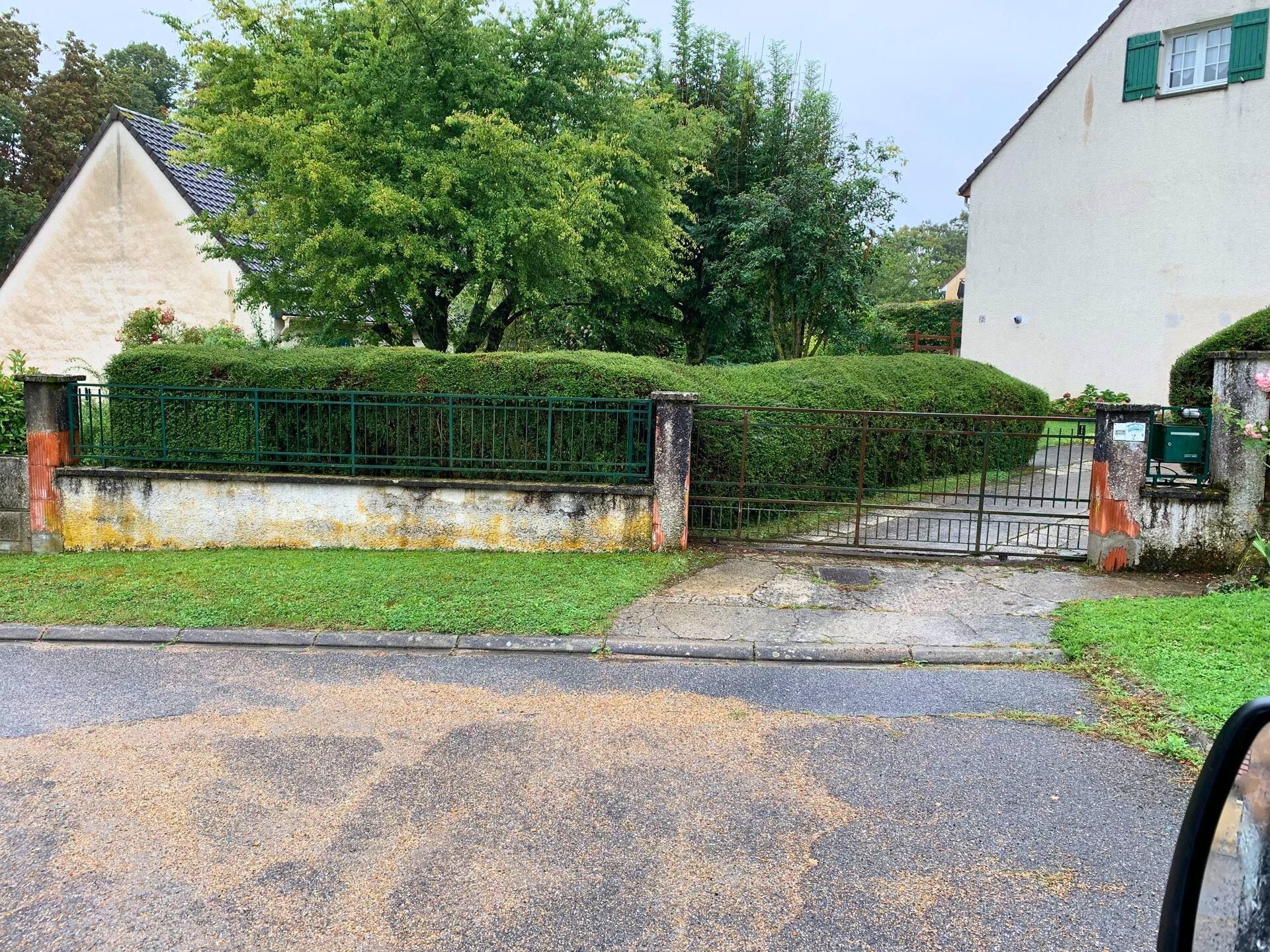 Une maison avec une allée privée fermée par un portail, des haies vertes et des arbres. La route est jonchée de petits débris.