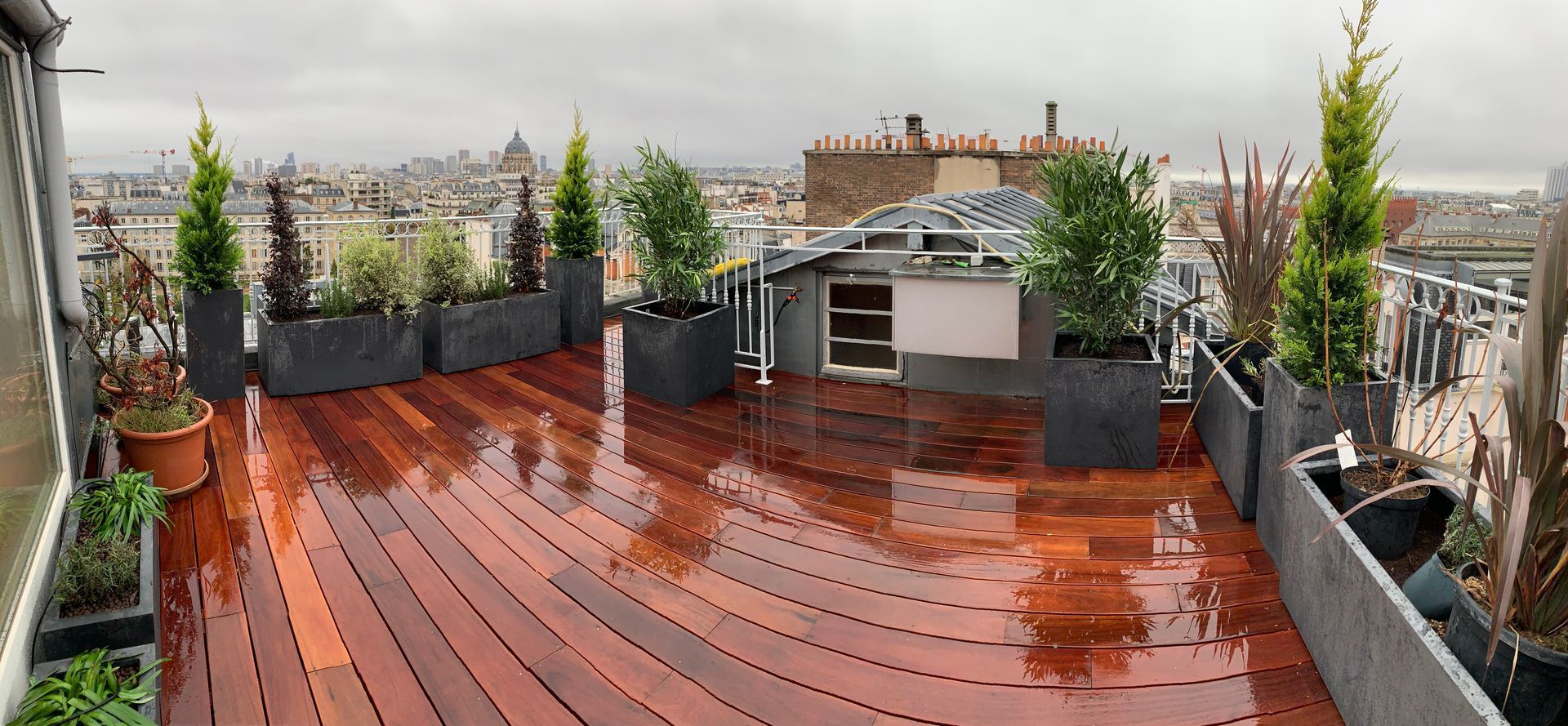 Terrasse sur le toit avec plancher en bois, jardinières et vue sur la ville sous un ciel nuageux.