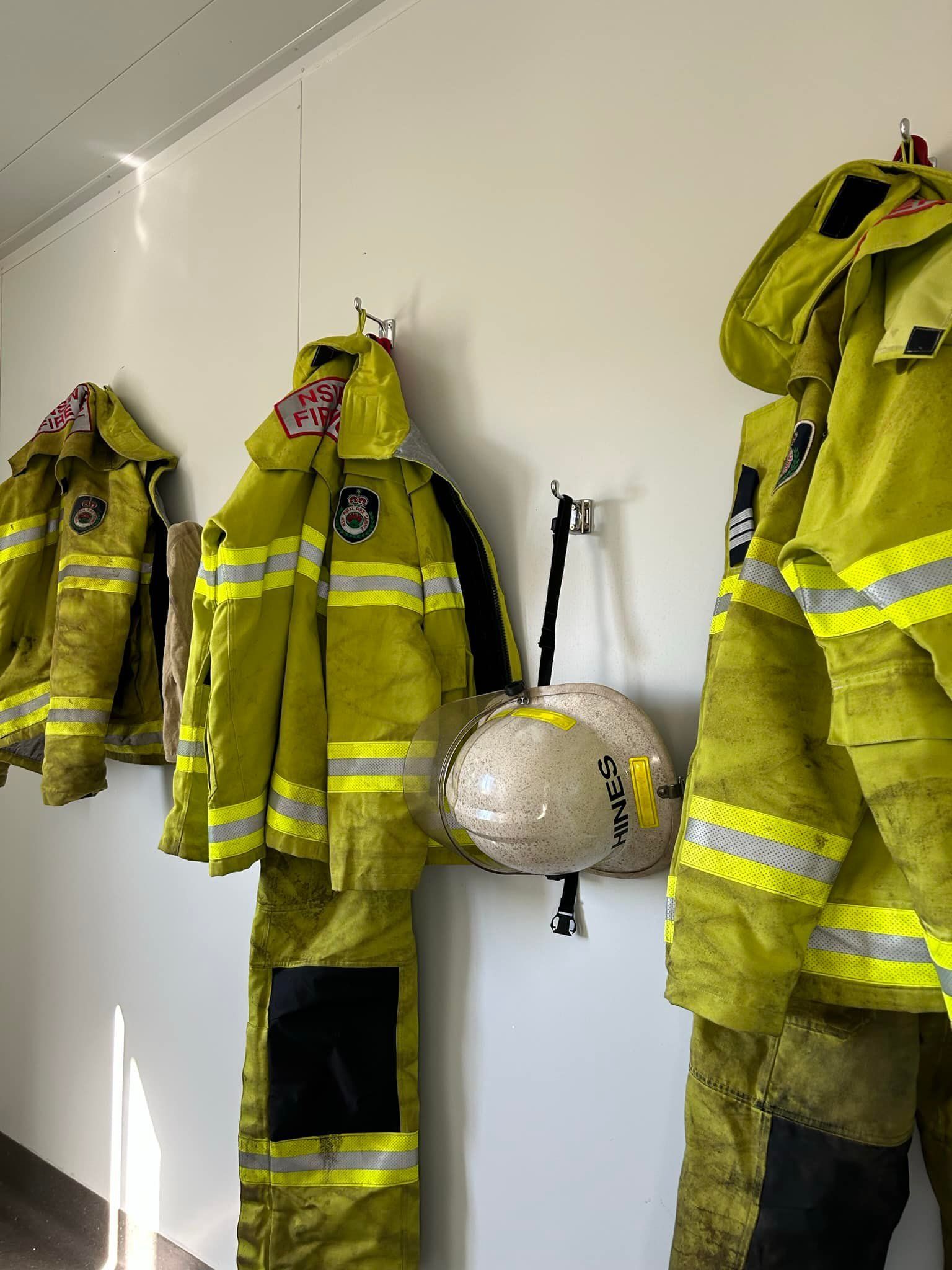 Yellow Firefighter Turnout Gear and a White Helmet Hanging on Hooks Against a Plain Wall — OSM Transportables In Dubbo, NSW