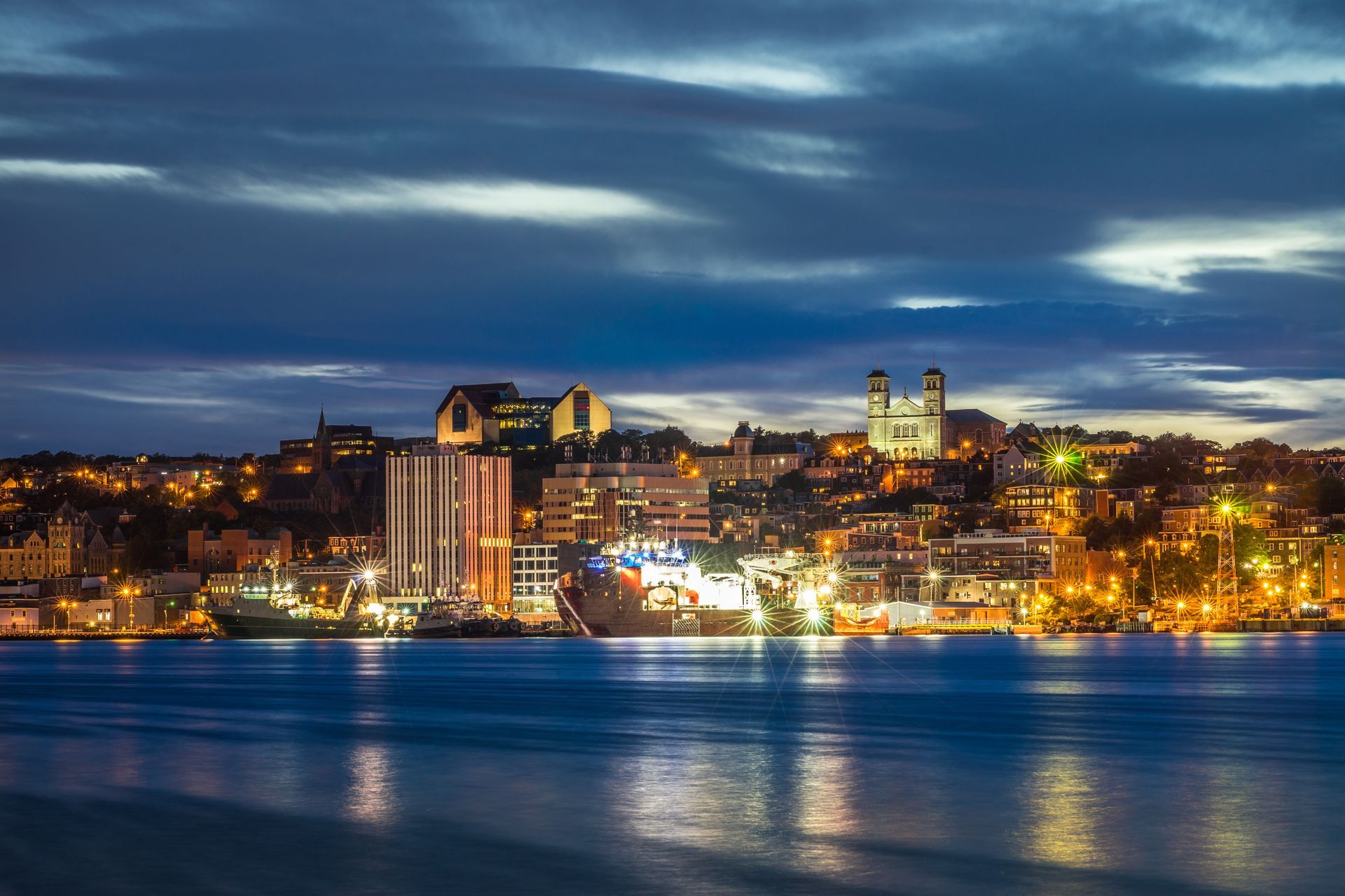 Night view of a city skyline with buildings illuminated along a dark blue harbor.