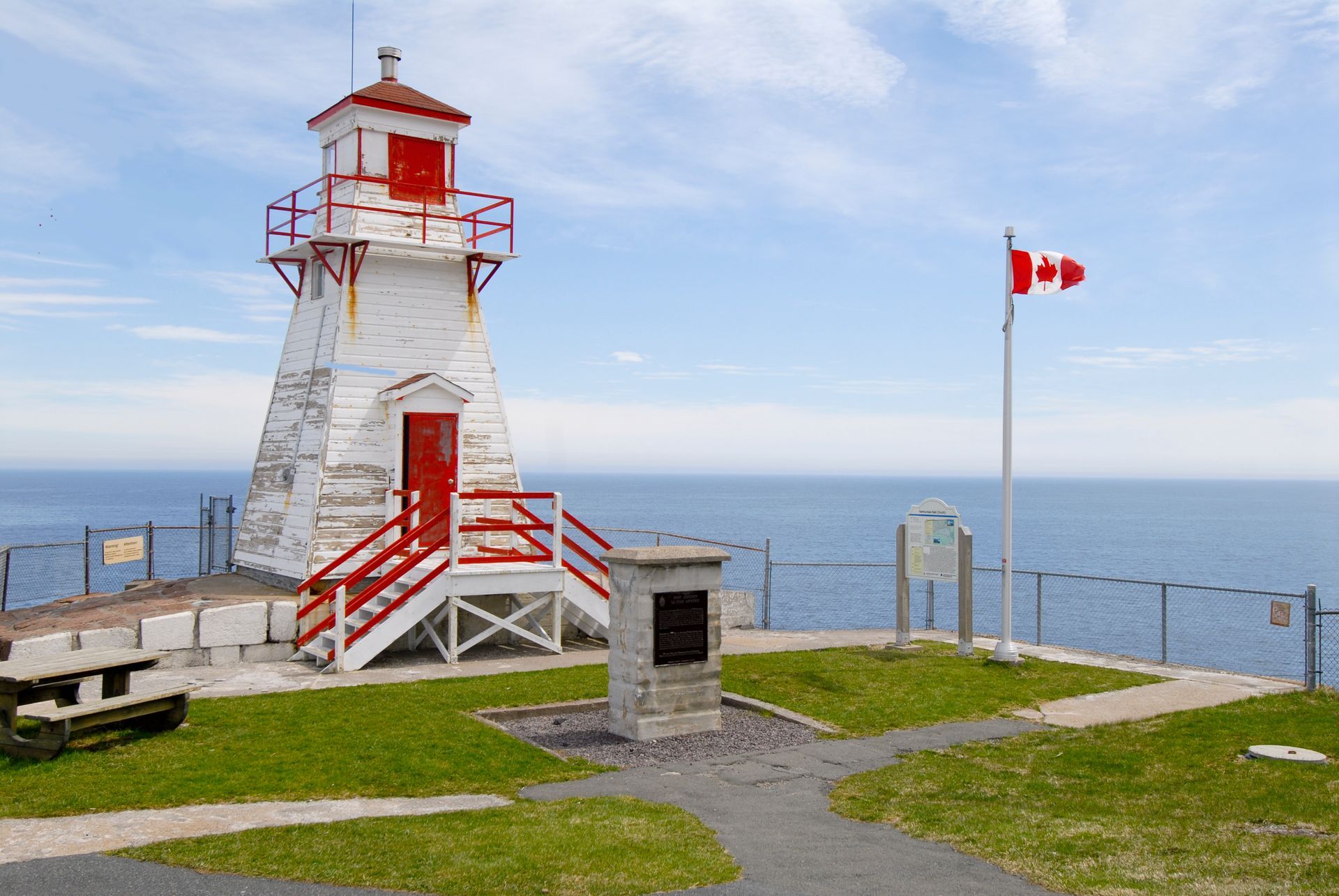 Lighthouse with red accents, Canadian flag, and monument on a grassy hill overlooking the ocean.