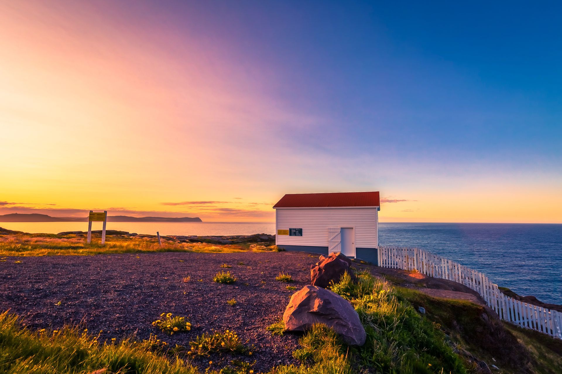 Small white building with red roof, overlooking the ocean at sunset; colorful sky.