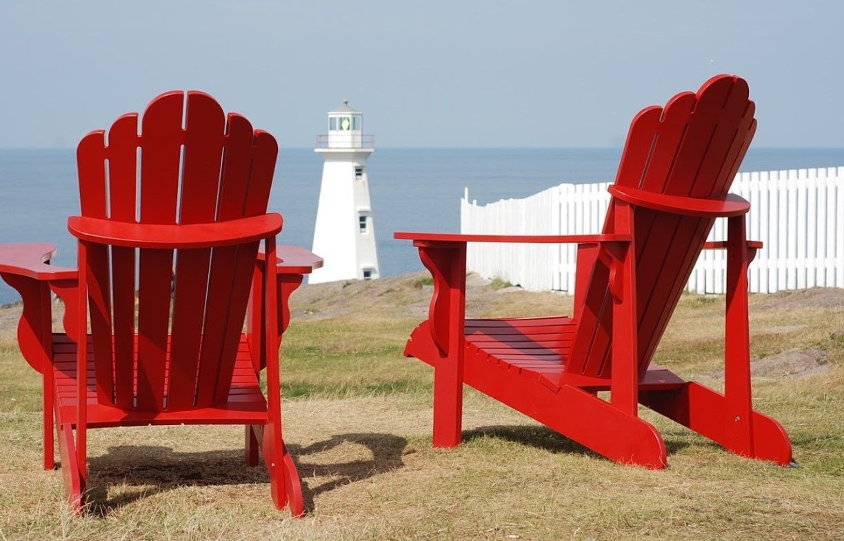 Two red Adirondack chairs face a white lighthouse and the ocean.