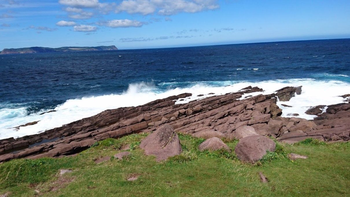 Ocean waves crashing on a rocky, brown shoreline under a blue sky with clouds.
