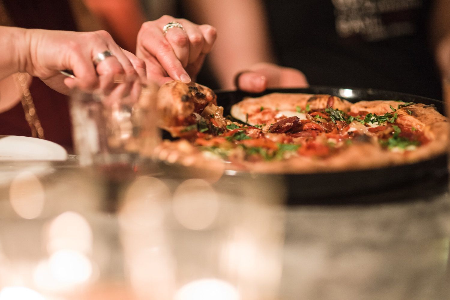 Hands cutting a slice of pizza from a round pan on a table, ready to be served.