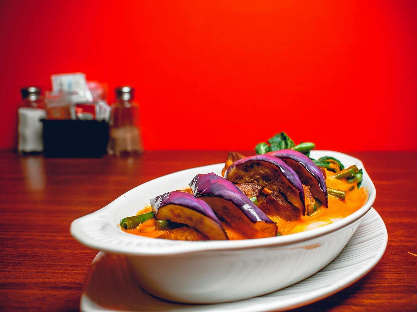Dish of eggplant in orange sauce, on a white plate, set on a wooden table, against a red backdrop.