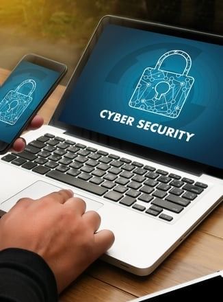 A person is typing on a laptop computer while holding a cell phone.