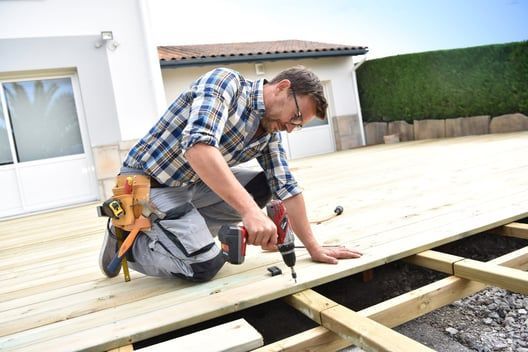 A man is kneeling down while using a drill on a wooden deck.