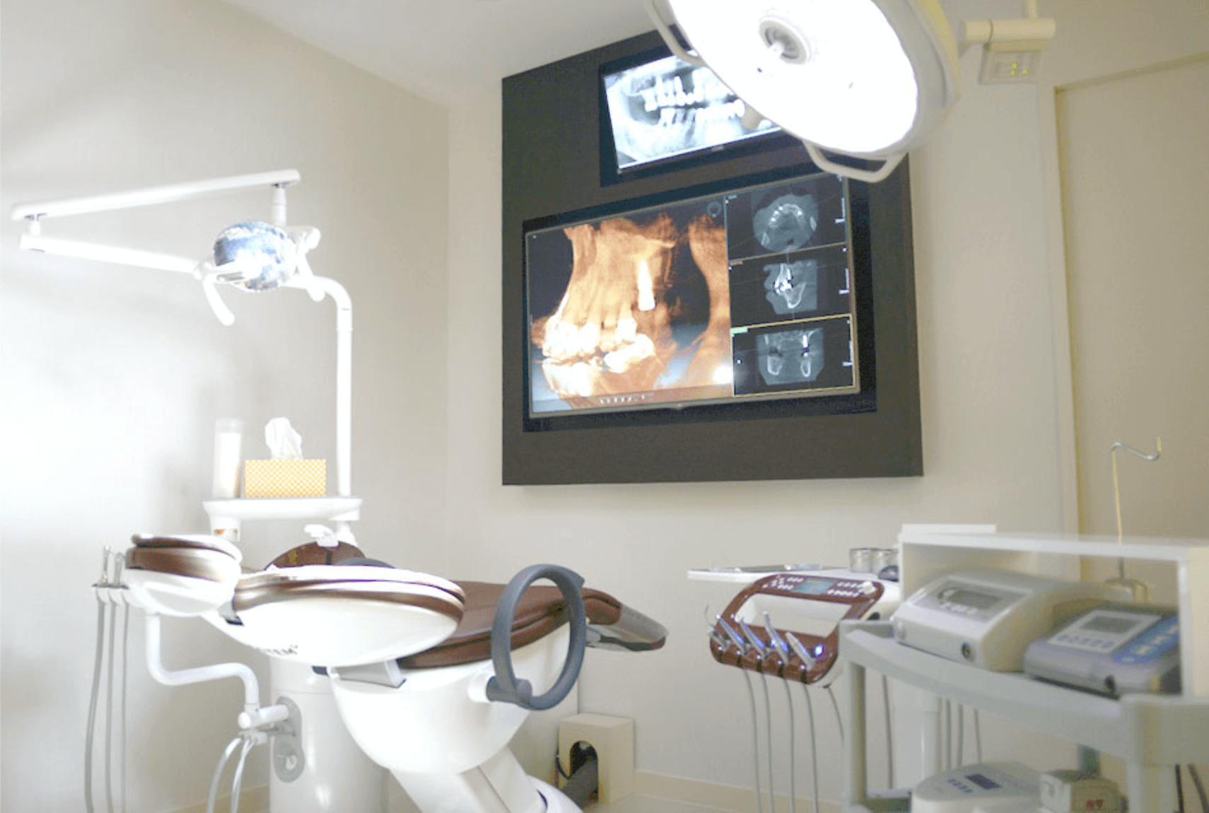 Dental chair in a bright office, with monitor displaying an X-ray and overhead lights.