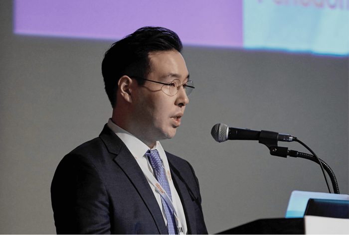 Man in suit speaking at a podium, wearing glasses, giving a presentation indoors.