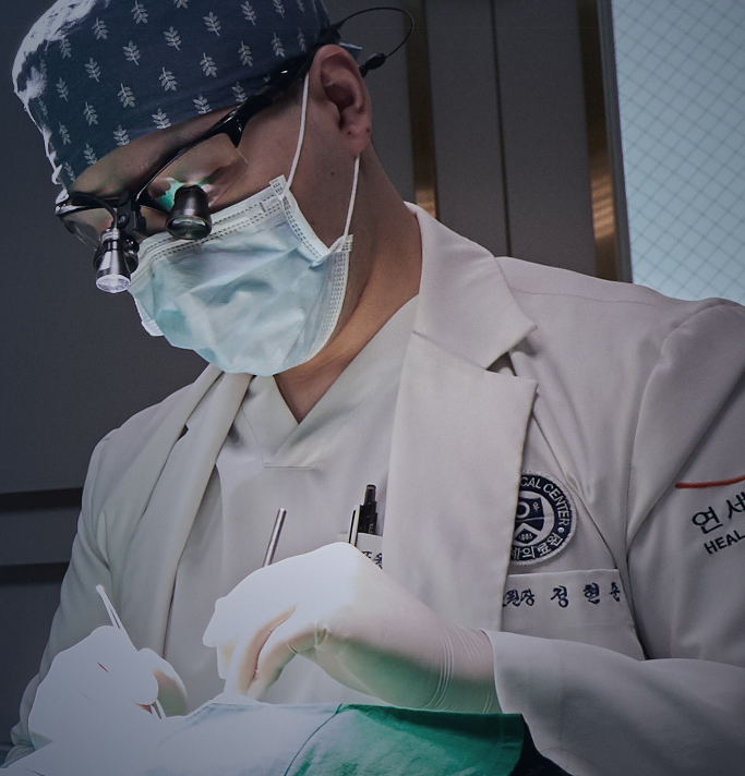 Surgeon in white coat and cap operating on a patient. He wears glasses, a mask, and surgical loupes.