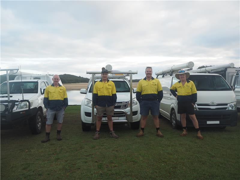 Four workers in work uniforms stand in front of their vehicles on grass, cloudy sky background— VR Marsden Plumbing in Red Rock, NSW