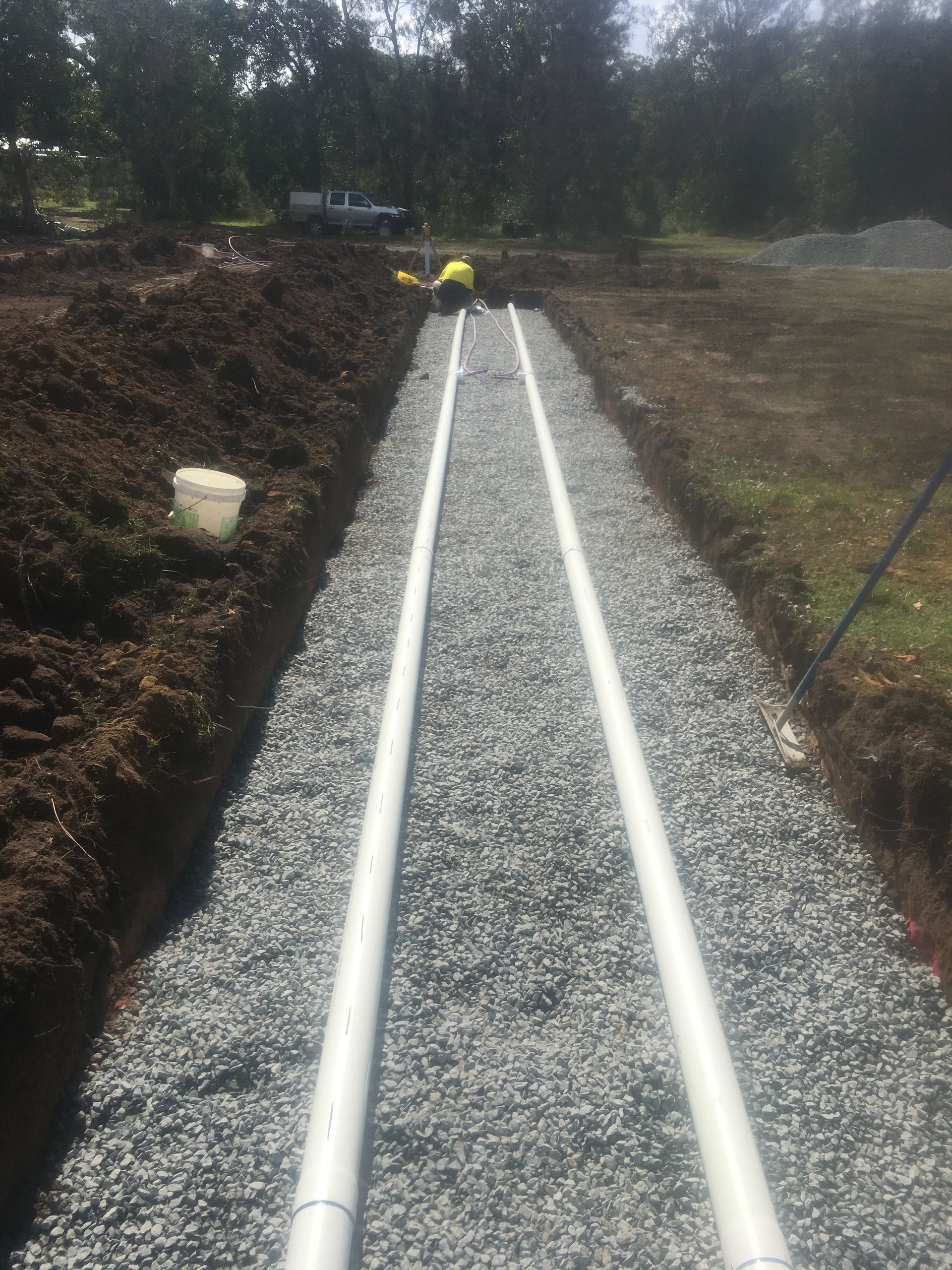 Trenched Gravel with Two Parallel White Pipes; a Worker in A Yellow Shirt — VR Marsden Plumbing in Red Rock, NSW