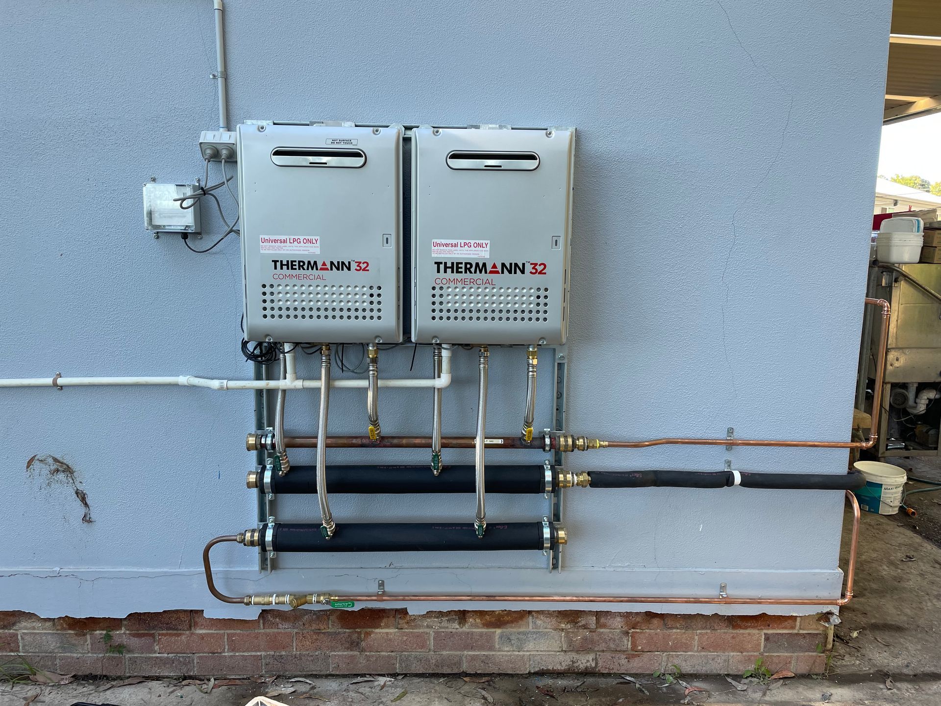 Two gray tankless water heaters mounted on a stucco wall with copper pipes and insulation. — VR Marsden Plumbing in Red Rock, NSW