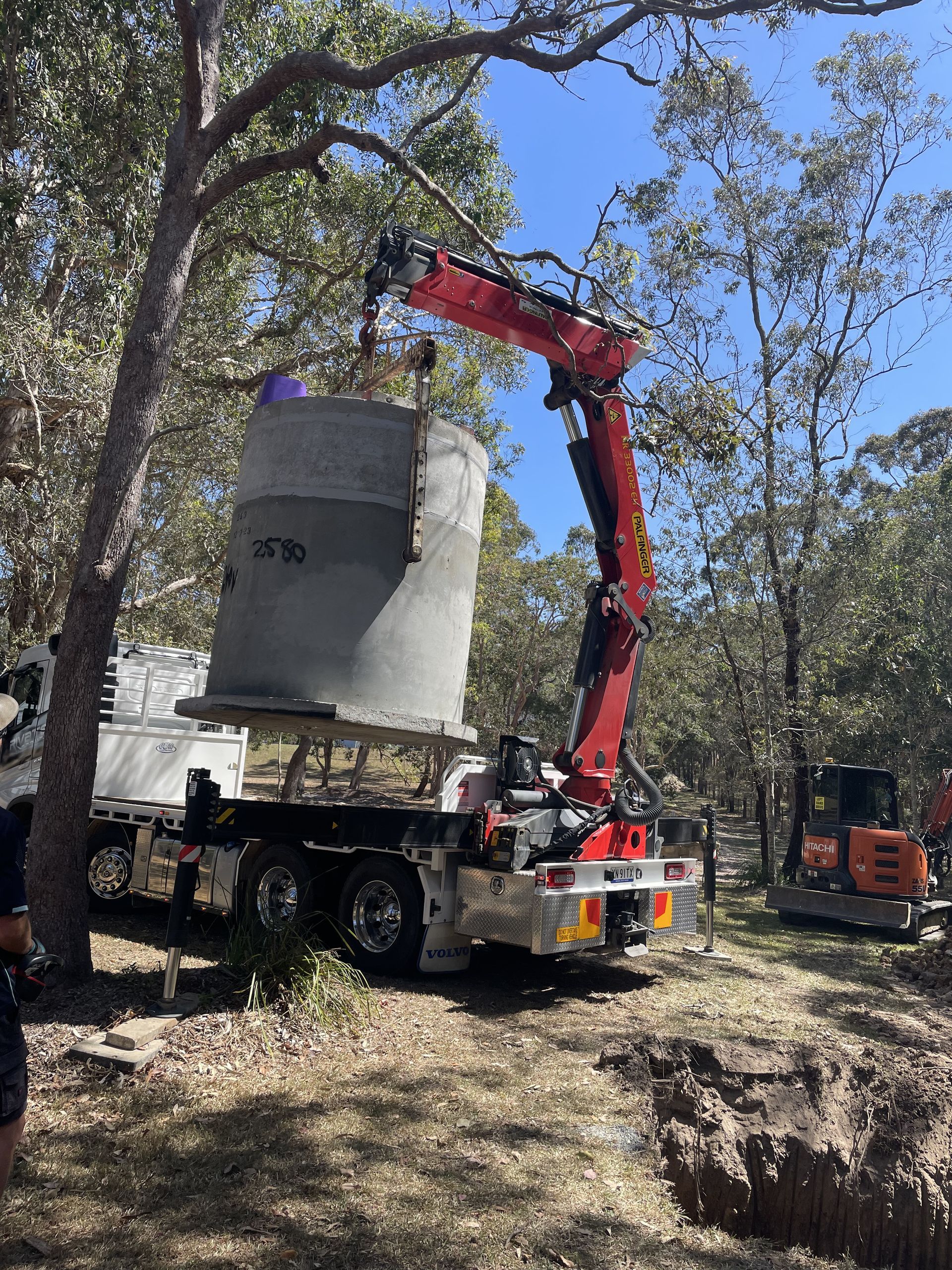 A truck with a crane lifting a large concrete cylinder in a wooded area under a blue sky— VR Marsden Plumbing in Red Rock, NSW