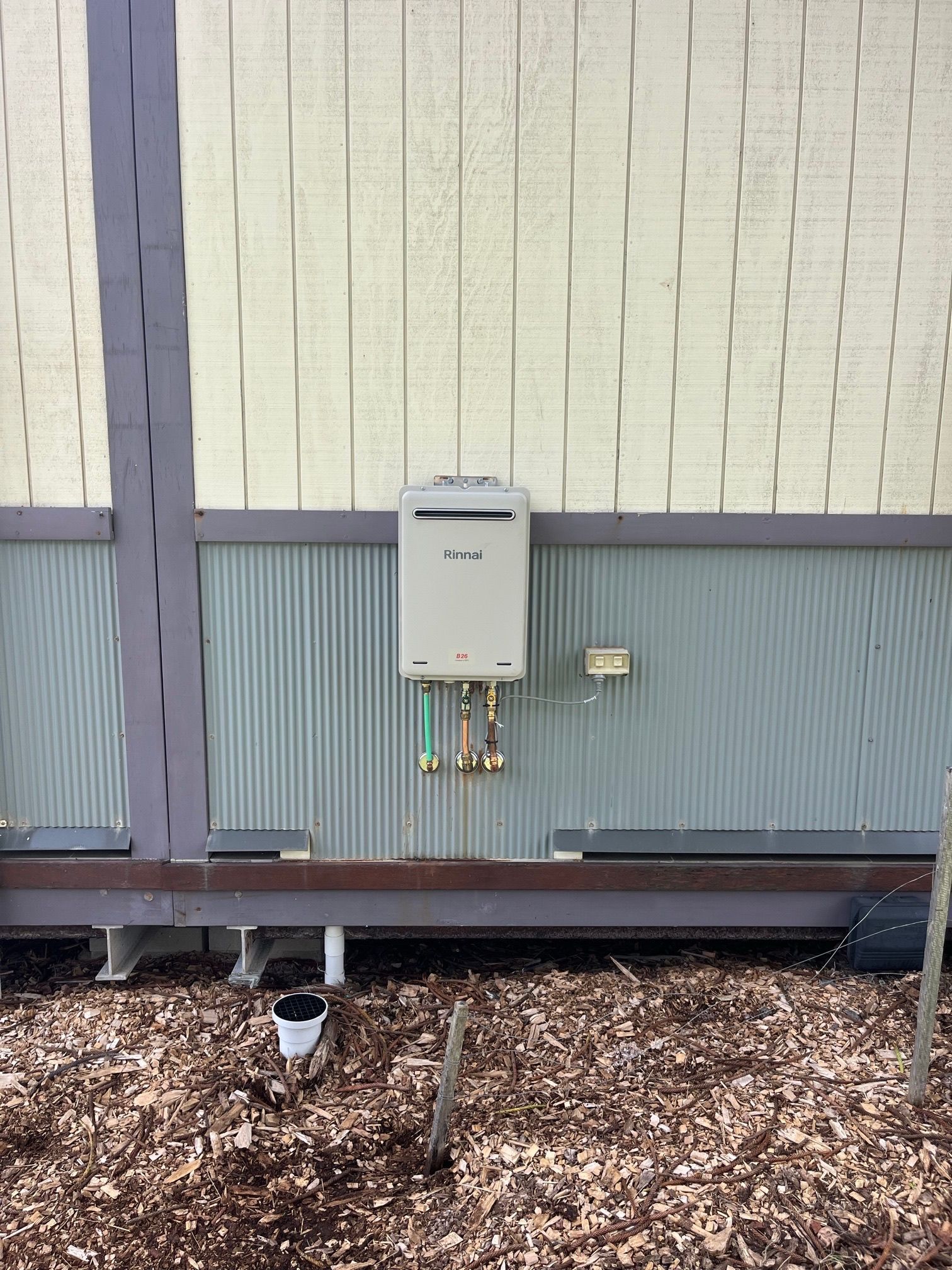 Water heater mounted on a wall with green and white siding. Below, brown mulch and a pipe. — VR Marsden Plumbing in Red Rock, NSW