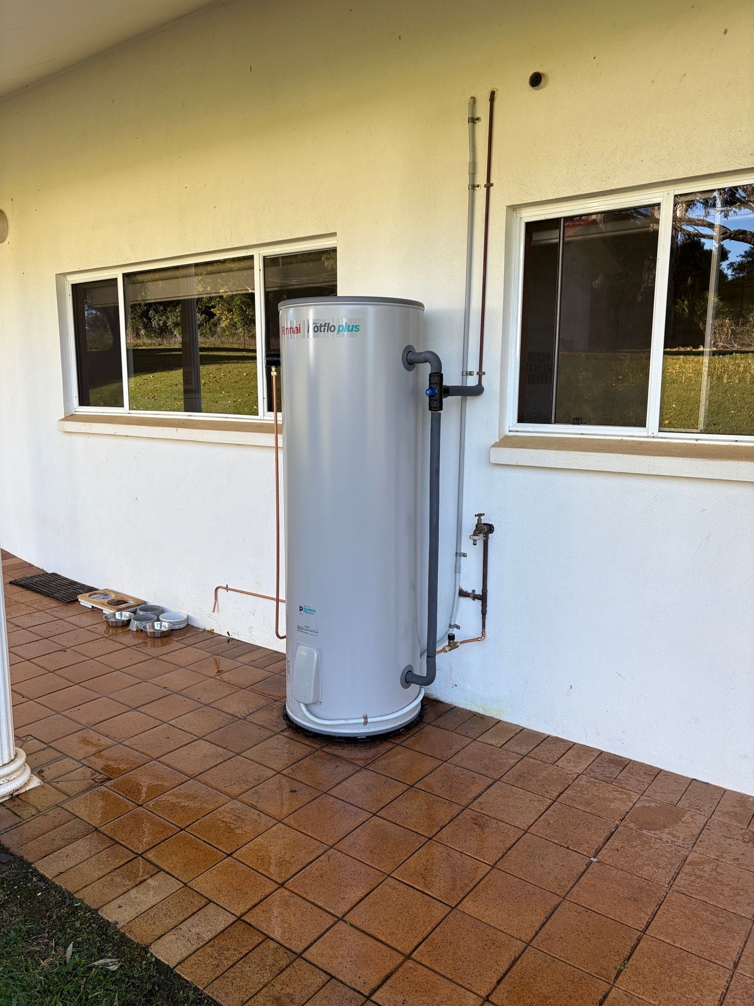 A water heater installed outside on a patio against a white wall with windows. — VR Marsden Plumbing in Red Rock, NSW