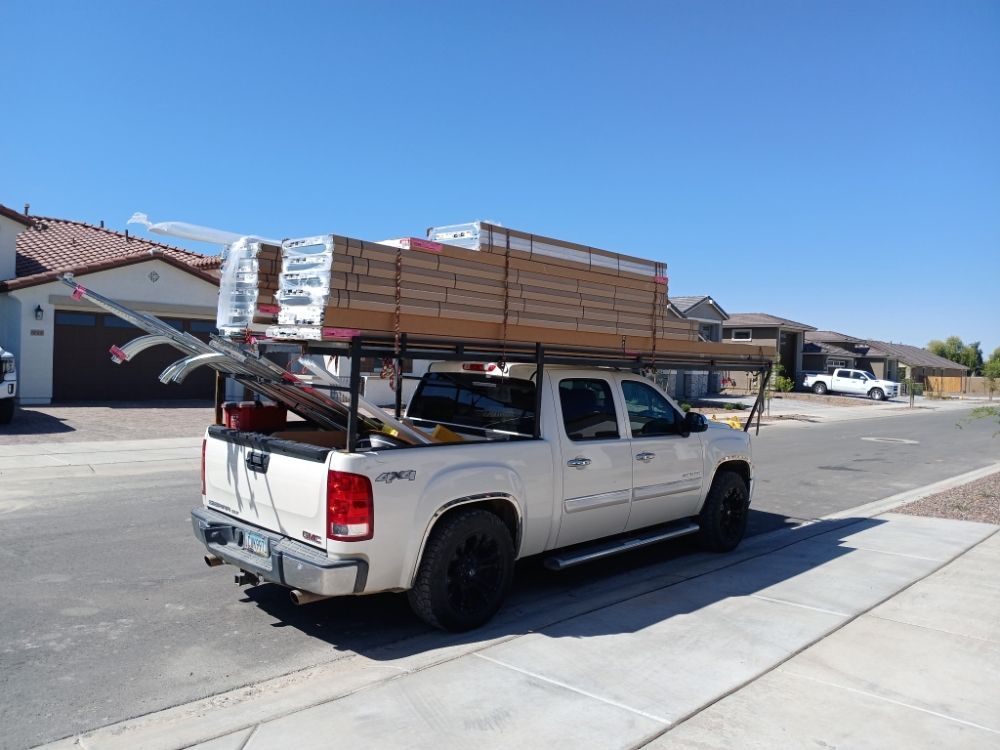 White pickup truck with a black rack carrying lumber, parked on a residential street under a sunny sky.