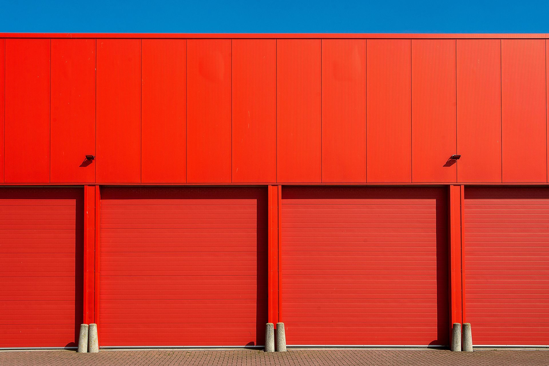 Red warehouse exterior with four closed roll-up doors and a blue sky.