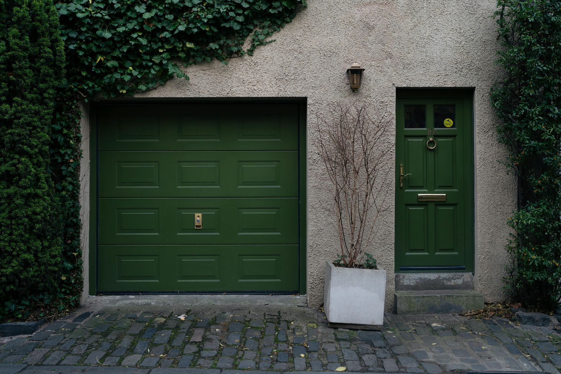 Green garage door and matching front door on a building with ivy, a cobblestone driveway, and a concrete planter.