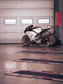 Motorcycle in a garage setting.  Black and white motorcycle on stand, in front of a closed garage door.