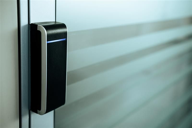 Black access card reader on a frosted glass door. A blue light is on at the top of the reader.