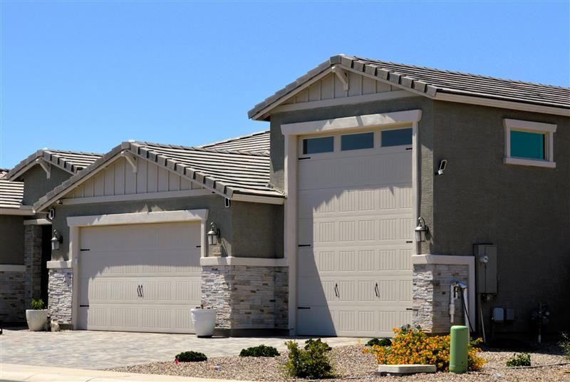 Exterior view of modern homes with light-colored stucco siding, beige garage doors, and stone accents against a blue sky.