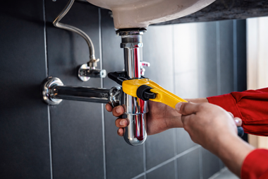 A person in red sleeves uses a yellow wrench to tighten a chrome pipe under a sink against a black tiled wall.