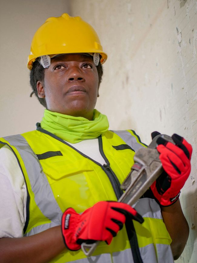 A worker in a yellow hard hat and high-visibility vest holds a pipe wrench while looking toward a wall.
