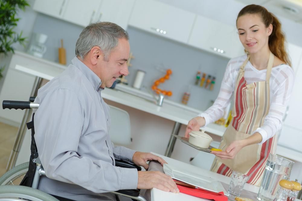Woman is Serving Food to a Man in a Wheelchair in a Kitchen — Serenity Strides in Redbank Plains, QLD