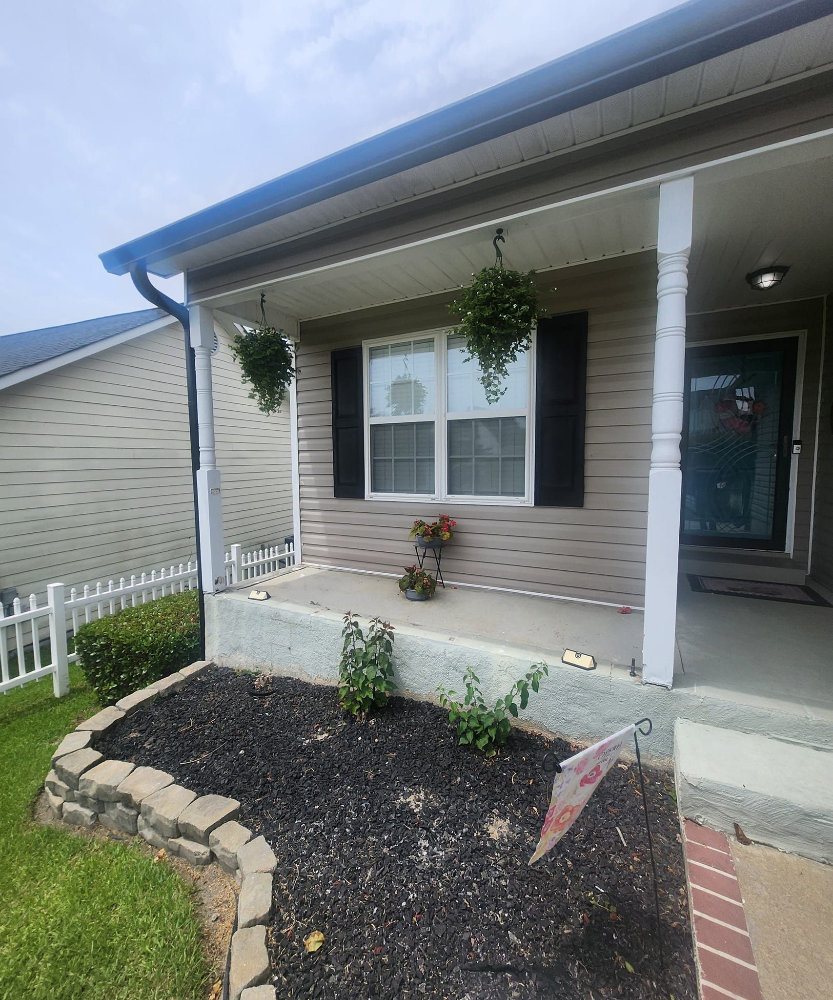 Front porch with hanging plants, black shutters, and a small flower bed with black mulch.