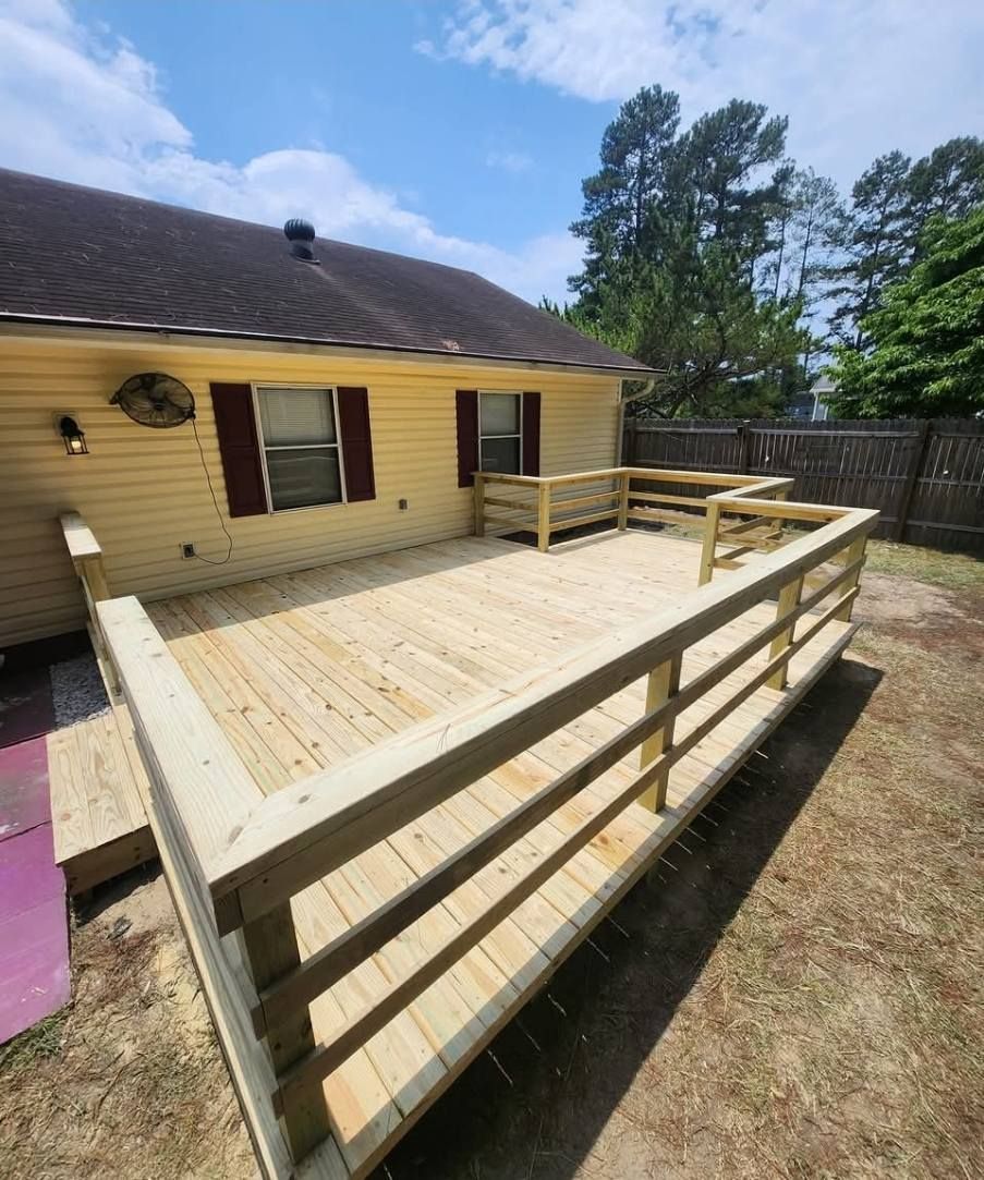 Wooden deck with railings attached to a yellow house with a dark roof, in a grassy backyard, on a sunny day.