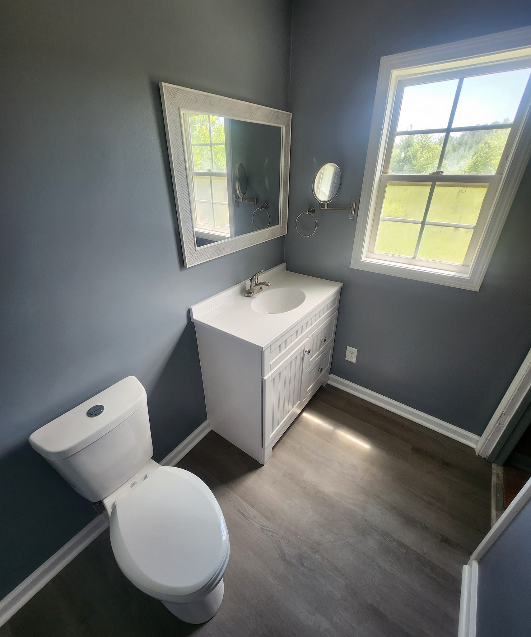 Bathroom with white vanity, mirror, toilet, and window on blue-gray walls.