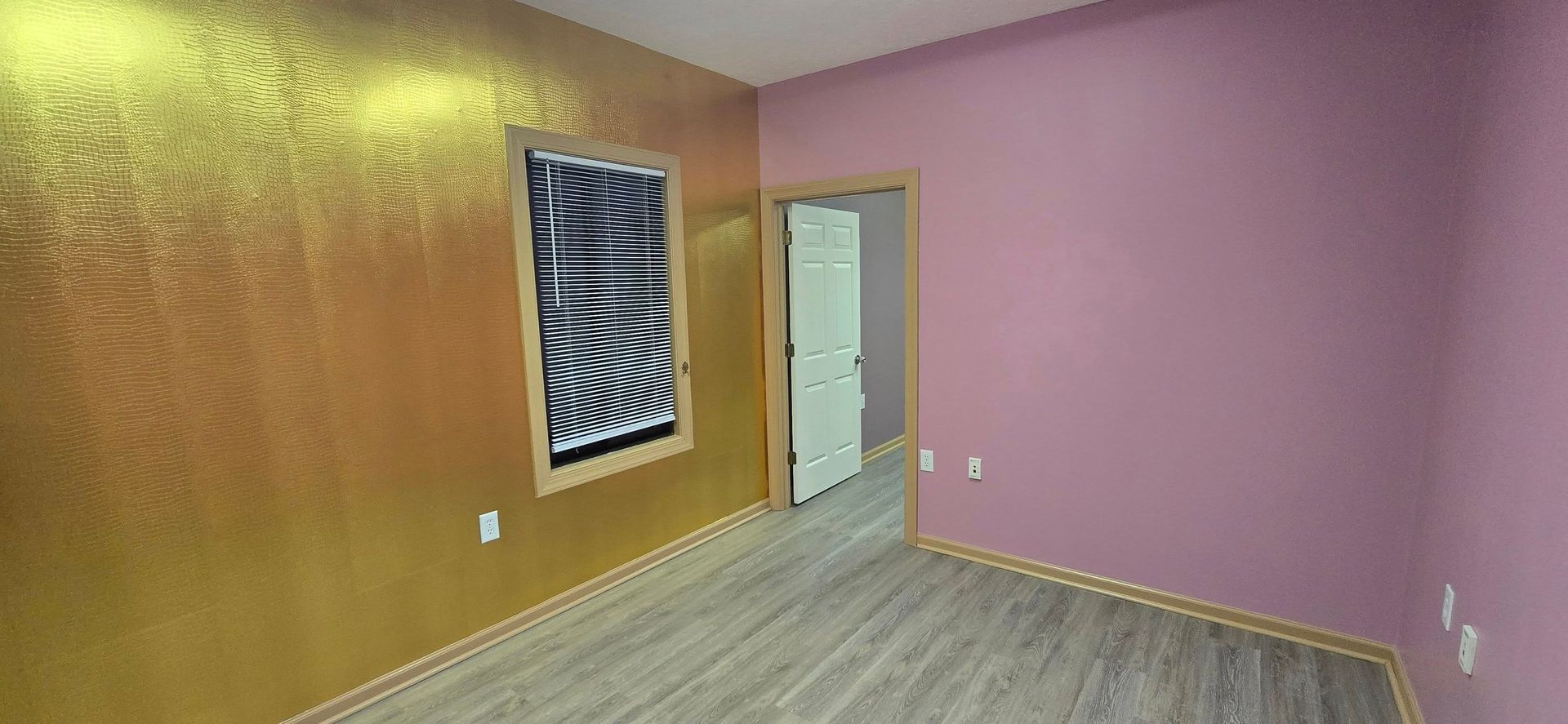 Interior room with gold textured wall, window, and pink wall with a door. Gray wood floors.